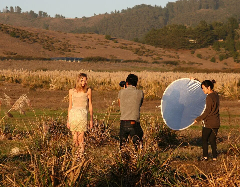 A person stands in a field while another person points a camera toward them and a third holds a circular reflector.