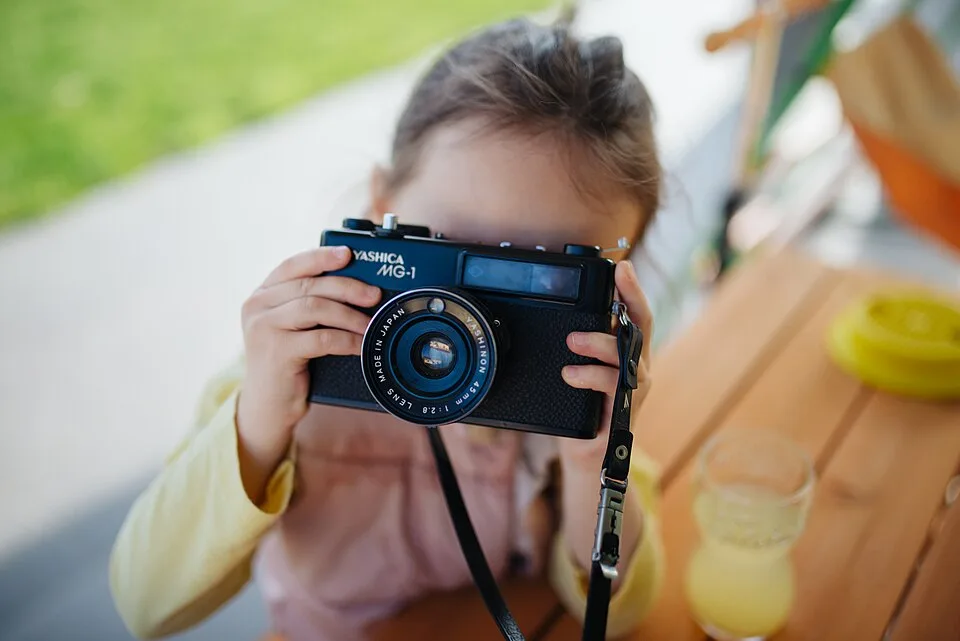 A child holds a black camera in front of their face while sitting beside a wooden table outdoors.