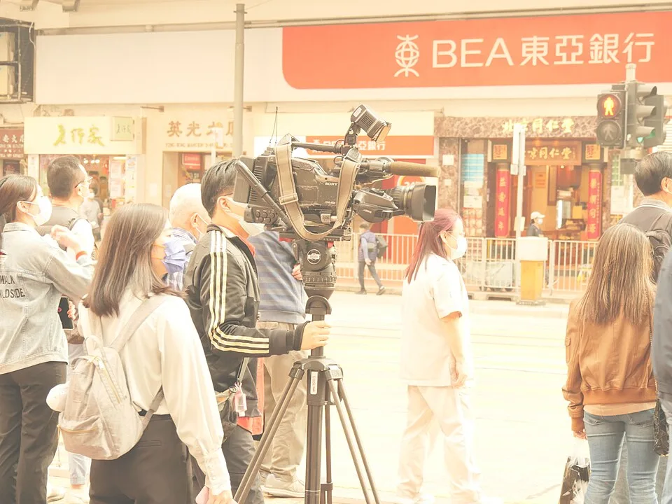 A camera operator stands beside a large video camera on a tripod among pedestrians on a city street.