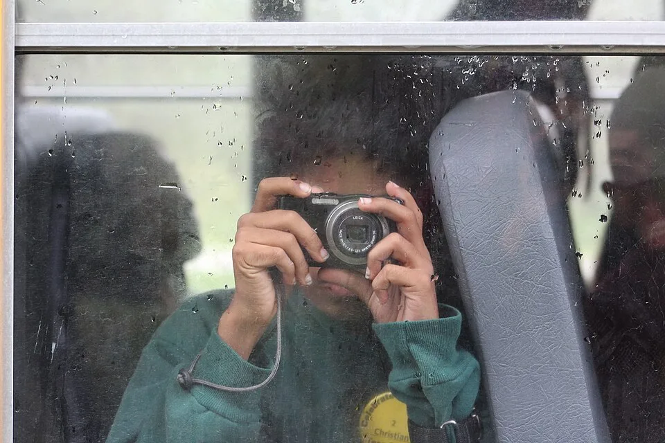A student holds a compact camera while seated behind a rain-spotted vehicle window.