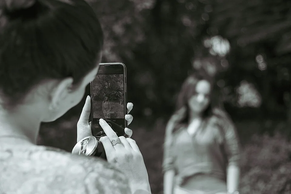 One person holds a phone toward another person standing outdoors; the phone screen displays a framed image.