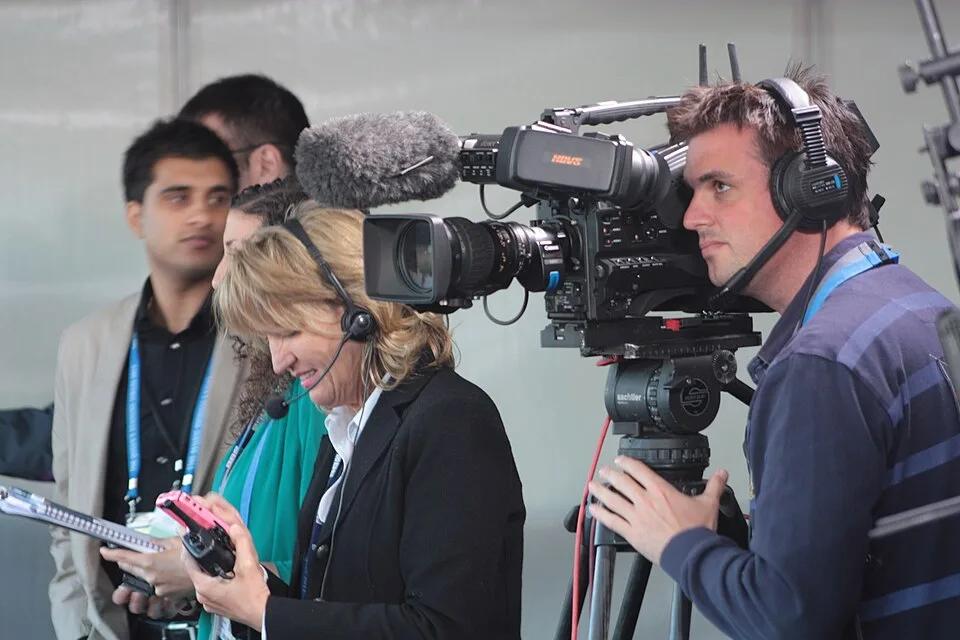 A camera operator wearing headphones stands beside a video camera while another person holds a device and notepad nearby.