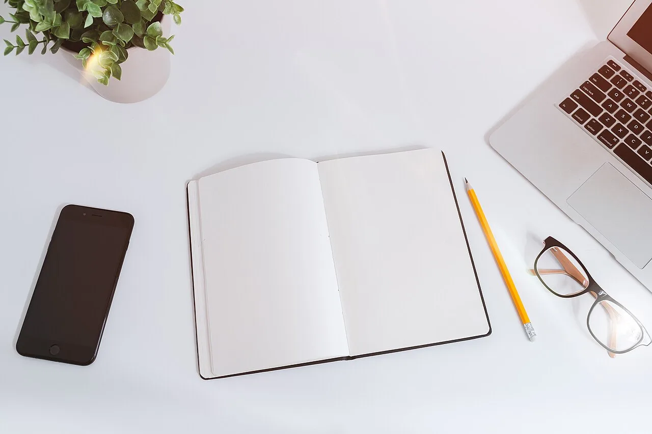 A phone, open notebook, pencil, glasses, and laptop sit on a white table.